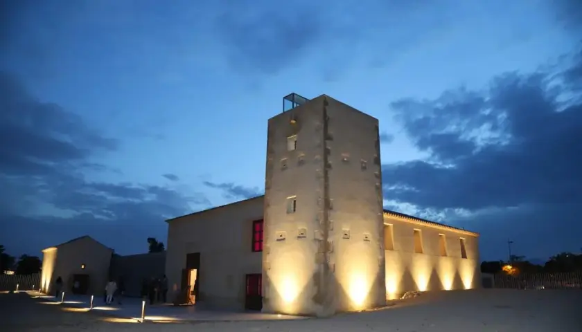 La torre Ansaldo, iluminada al anochecer, se alza junto a edificios bajos bajo un cielo azul oscuro con nubes.