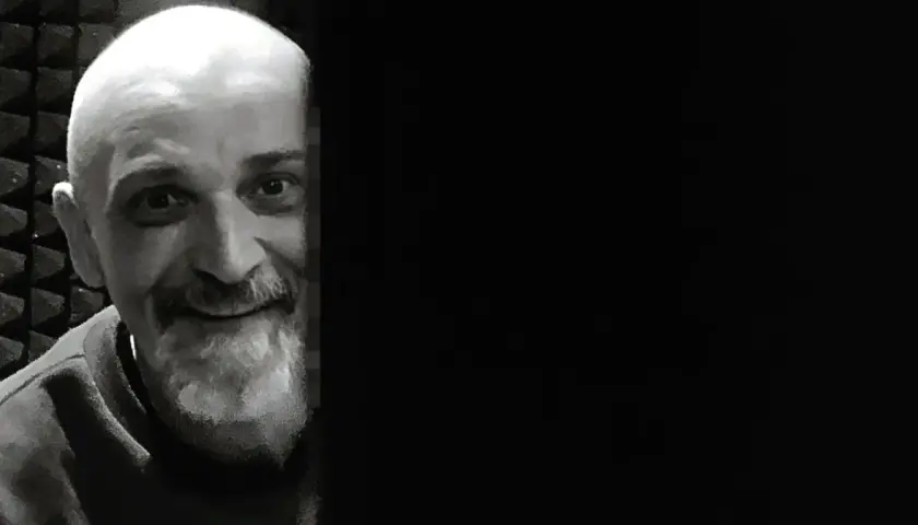 Uśmiechnięty Wojciech Sęp Dudziński, z siwą brodą i łysą głową, wygląda zza ciemnej zasłony.