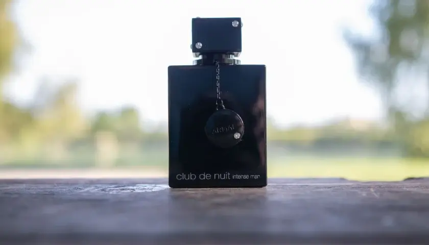 Elegancka czarna butelka perfum Club de Nuit Intense Man, ozdobiona łańcuszkiem i dyskiem z logo, stoi na drewnianym stole.