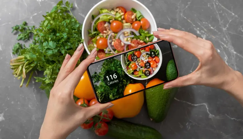 Ręce trzymają smartfon z aplikacją pokazującą kalorie. Na ekranie widać sałatkę i zioła, a obok na blacie leżą warzywa. Zastanawiasz się, ile kalorii żeby przytyć?