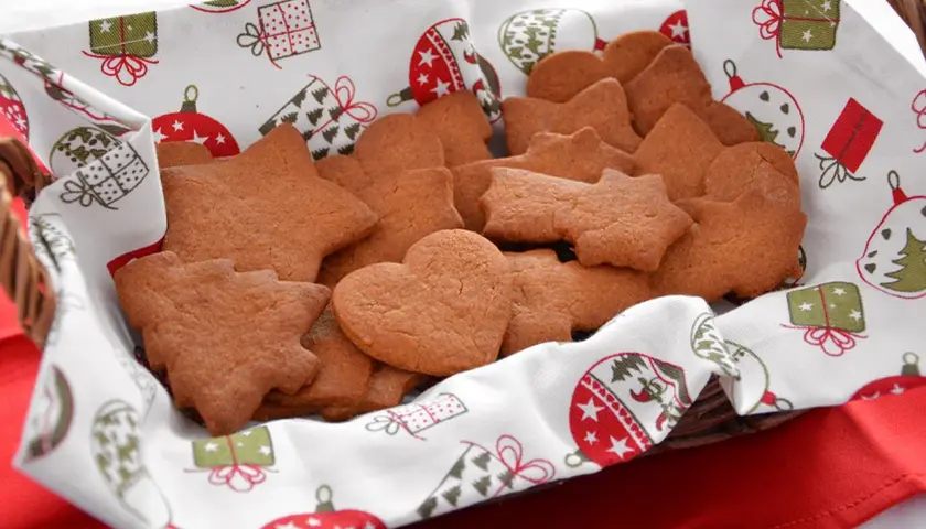Cesto pieno di biscotti alla cannella a forma di stella, cuore e albero, su un telo natalizio.