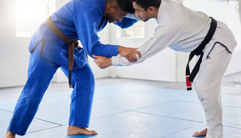 Dwóch mężczyzn w kimonach do BJJ, jeden w niebieskim z brązowym pasem, drugi w białym z czarnym pasem, ćwiczy chwyty na macie.