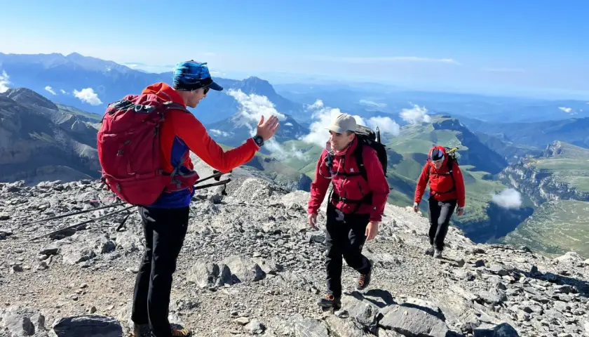 Tres excursionistas suben por una cresta rocosa en el **alt Pirineu**. El sol brilla en un cielo azul claro, con vistas a un vasto paisaje montañoso.