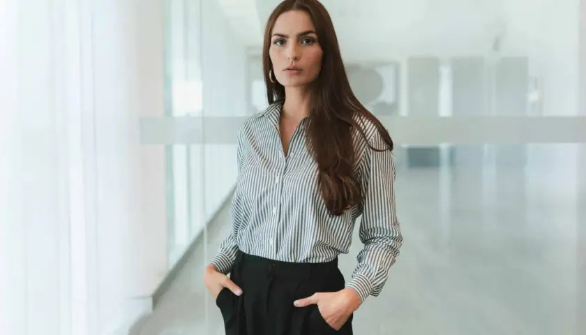 Frau in gestreifter Bluse und schwarzen Hosen, ein Look im business casual Stil, steht mit den Händen in den Taschen.