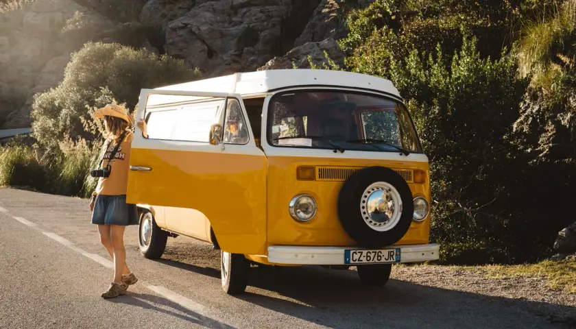 A woman with a straw hat and camera leans against a yellow van, ready for an adventure in Majorque en van.