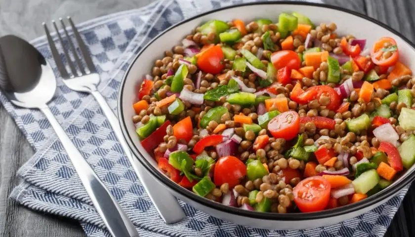 Un'insalata di lenticchie colorata e fresca, con pomodorini, peperoni, cipolla rossa e carote. Servita in una ciotola bianca, accanto a posate d'argento su un tovagliolo a quadri blu.