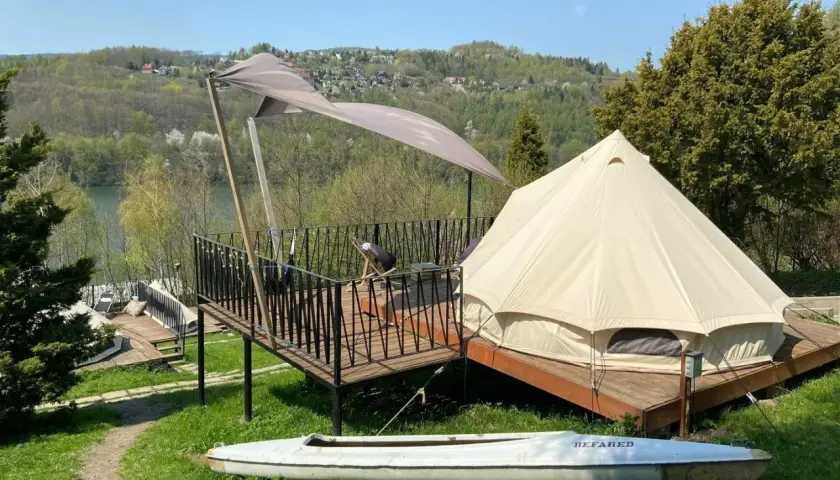 Namiot typu glamping camp z widokiem na jezioro i wzgórza. Na tarasie stolik i krzesło. Obok leży kajak.