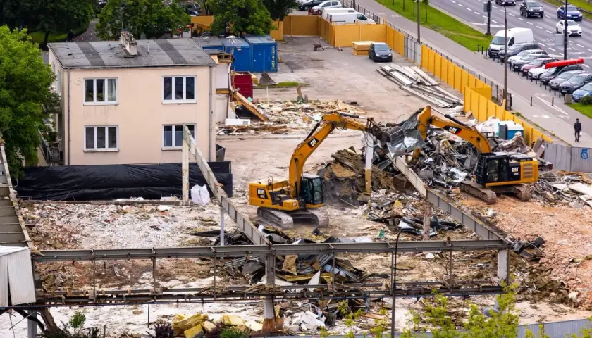 Dwa koparki pracują na placu budowy, rozbierając budynek. W tle widać drogę z samochodami i drzewa.