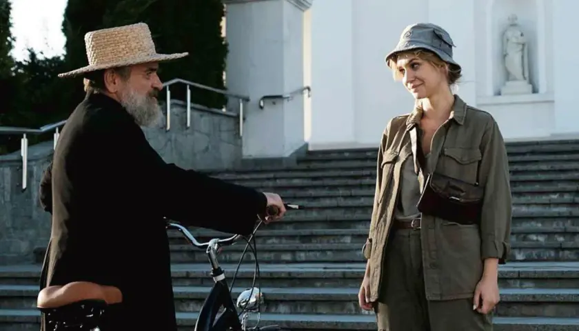 Scena z filmu Jacka Bromskiego: mężczyzna w słomkowym kapeluszu i czarnej sutannie przy rowerze rozmawia z kobietą w wojskowym kapeluszu.
