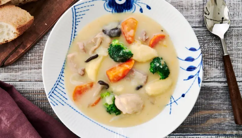 A hearty bowl of Japanese cream stew, brimming with chicken, carrots, potatoes, broccoli, and mushrooms, served with crusty bread.