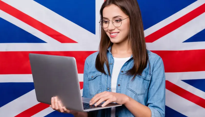 Uśmiechnięta dziewczyna w okularach pracuje na laptopie na tle flagi Wielkiej Brytanii, tworząc przykładowy artykuł po angielsku.