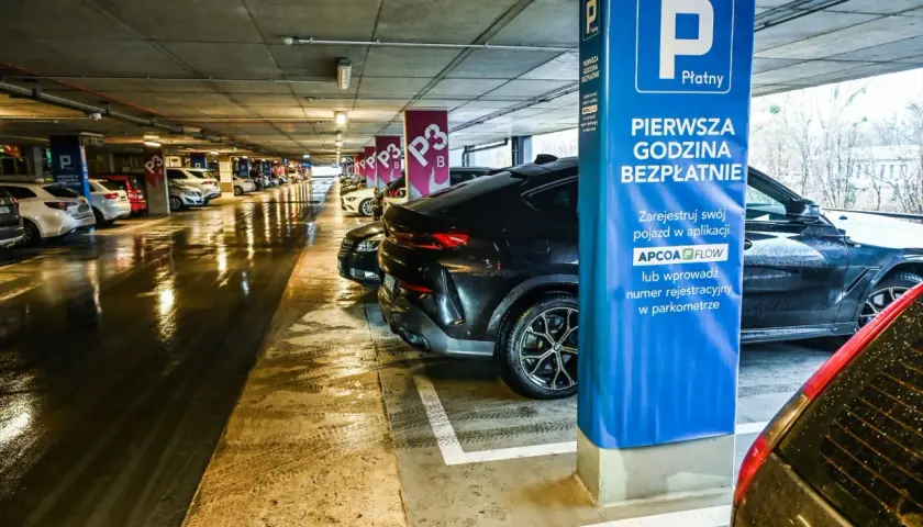 Mokry parking nad zaporą, rząd samochodów pod betonowym stropem. Niebieska tablica informuje o pierwszej darmowej godzinie.