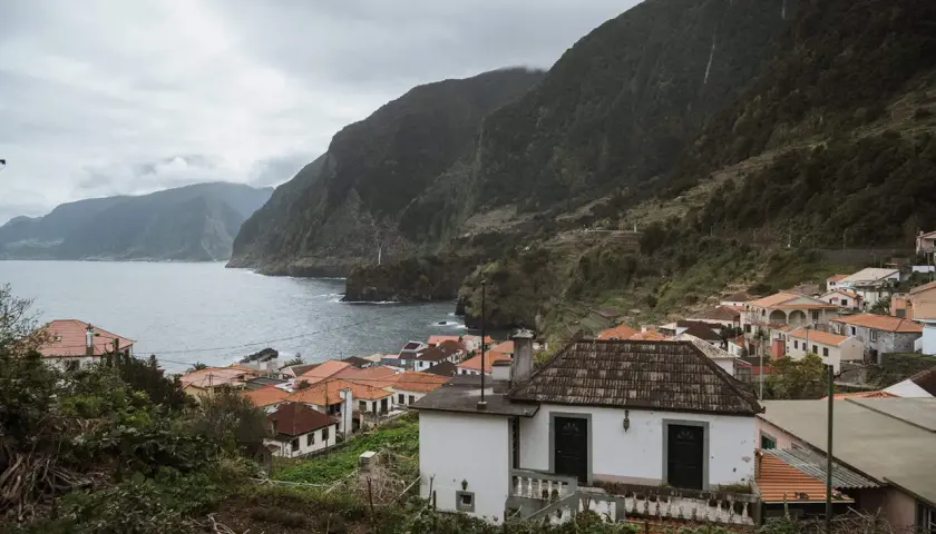 Coastal village with red-tiled roofs nestled against lush green mountains. A perfect place to stay in Madeira, offering stunning ocean views.