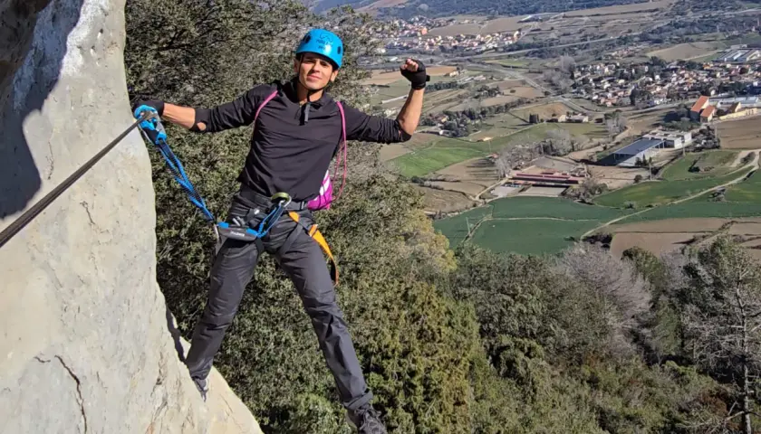 Hombre con casco azul y arnés, asegurado a un cable de **material vía ferrata**, disfruta de las vistas del valle.
