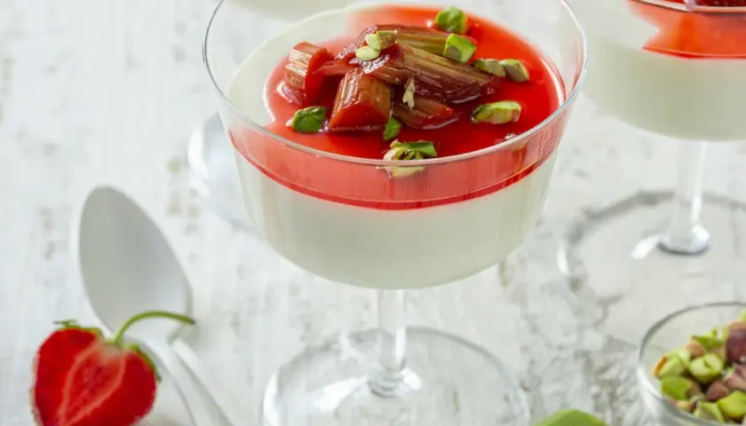 Panna cotta z rabarbarem i pistacjami w kieliszku. Słodki deser z rabarbarem, ozdobiony zielonymi orzeszkami.