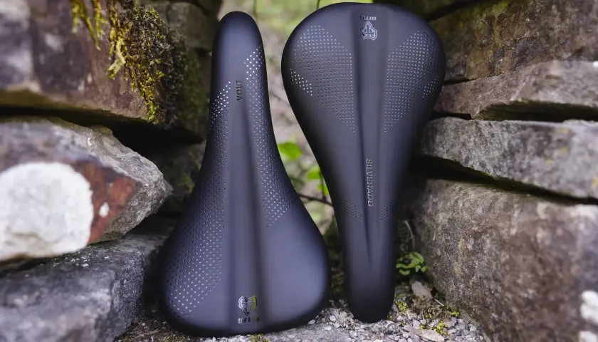 Two black WTB bike saddles, a Volt and a Silverado, rest against a stone wall, ready for adventure.