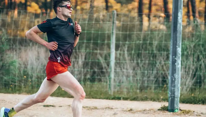 Mężczyzna w sportowym stroju biegnie, ćwicząc jak zwiekszyc kadencje w bieganiu. W tle siatka i drzewa.