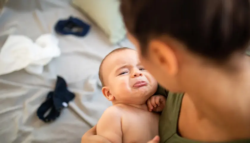 Un bebé con cara de llanto es sostenido por su madre. Parece que el bebe se cae de la cama y está asustado.