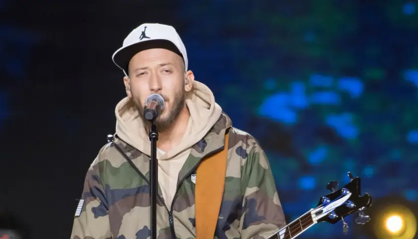 Artysta w czapce z daszkiem i kurtce moro śpiewa do mikrofonu, trzymając gitarę. Może to jego żona czeka na niego po koncercie?