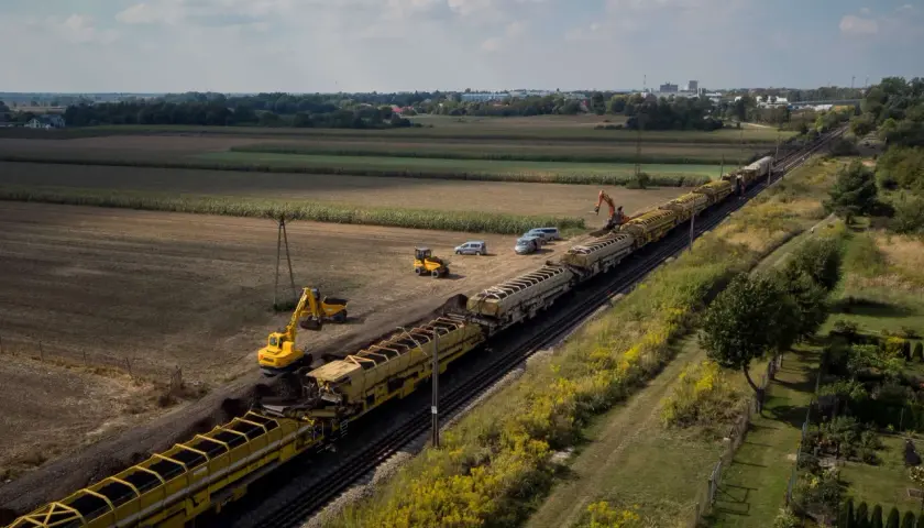 Długi pociąg z wagonami wypełnionymi ziemią, obok koparki i samochodów. Prace torowe w tle pól i zabudowań, widać odbojnica kolejowa.