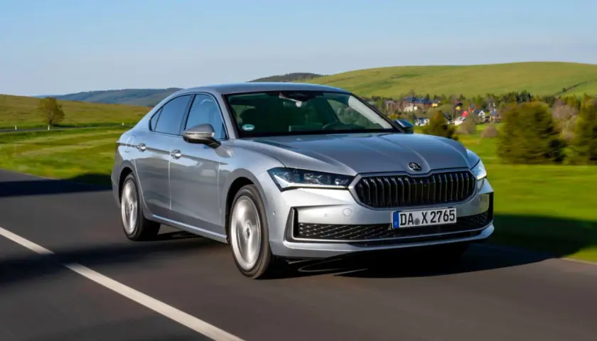 Srebrna, nowa Skoda Superb pędzi po drodze, mijając malownicze, zielone wzgórza i domki.