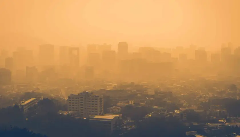 Vista aérea de una ciudad envuelta en una densa neblina anaranjada, con siluetas de edificios apenas visibles. Las partículas PM10 flotan en el aire, creando esta atmósfera opresiva.