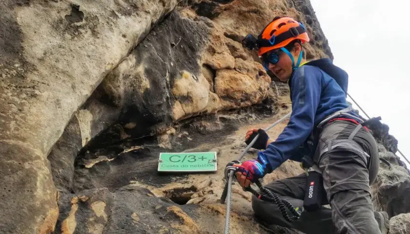 Młoda osoba w kasku wspina się po skale na via ferrata w Decinie. Tabliczka "Cesta do nebíčka" wskazuje trudność trasy.