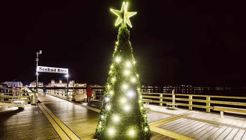 Ein festlich beleuchteter Weihnachtsbaum mit Sternspitze auf dem Holzsteg des Seebads Binz. Im Hintergrund die Lichter der Stadt.