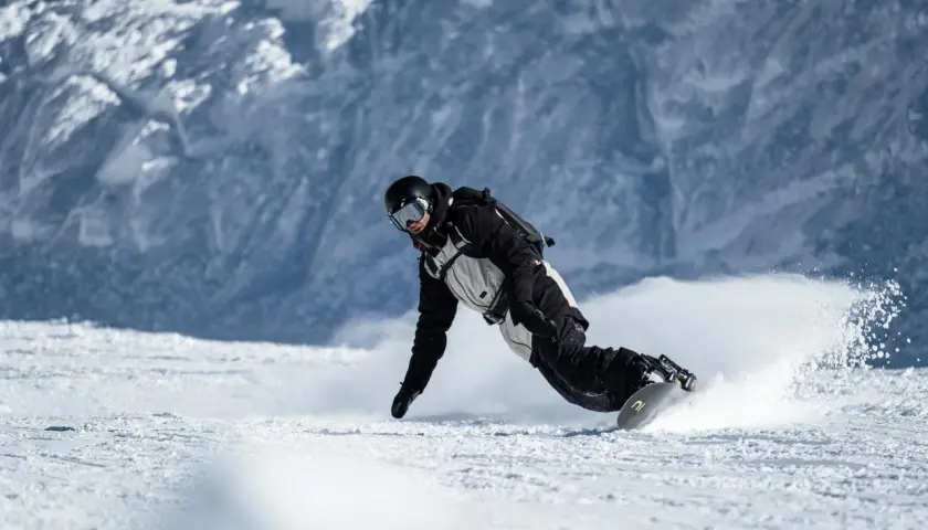 Snowboarder w kasku i goglach, z ochraniaczem nadgarstka snowboard, pochyla się nisko, tworząc obłok śniegu podczas zjazdu.