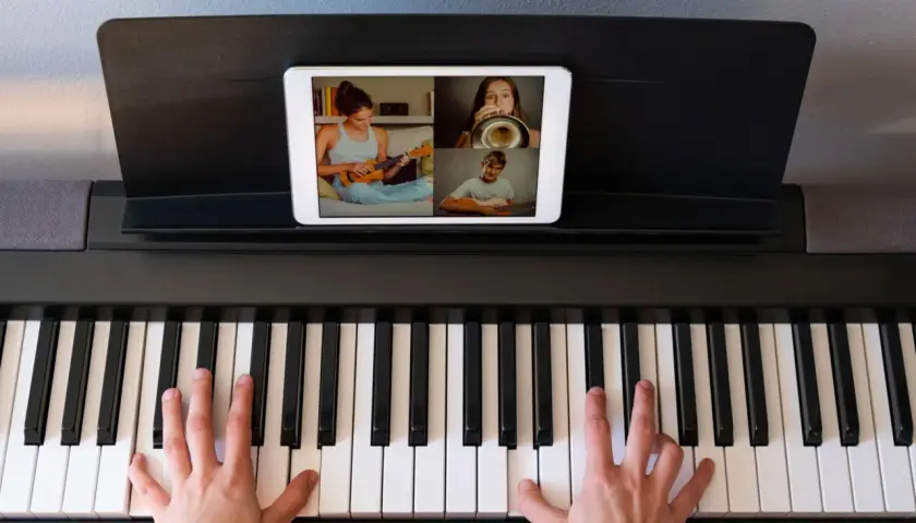 Ręce na klawiaturze pianina cyfrowego, na ekranie tabletu widać lekcję muzyki online z uczniami grającymi na ukulele, trąbce i gitarze.