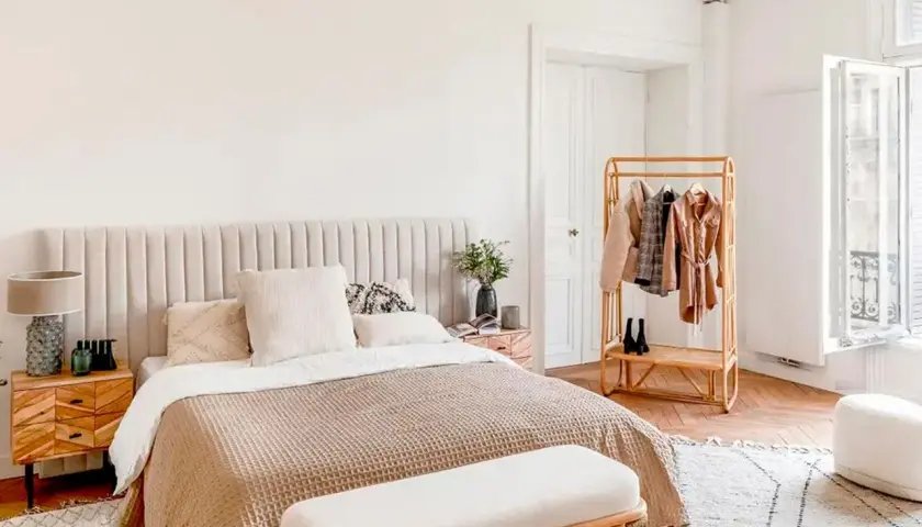 Chambre bohème scandinave avec lit douillet, table de chevet en bois, portant à vêtements en rotin et fenêtre lumineuse.