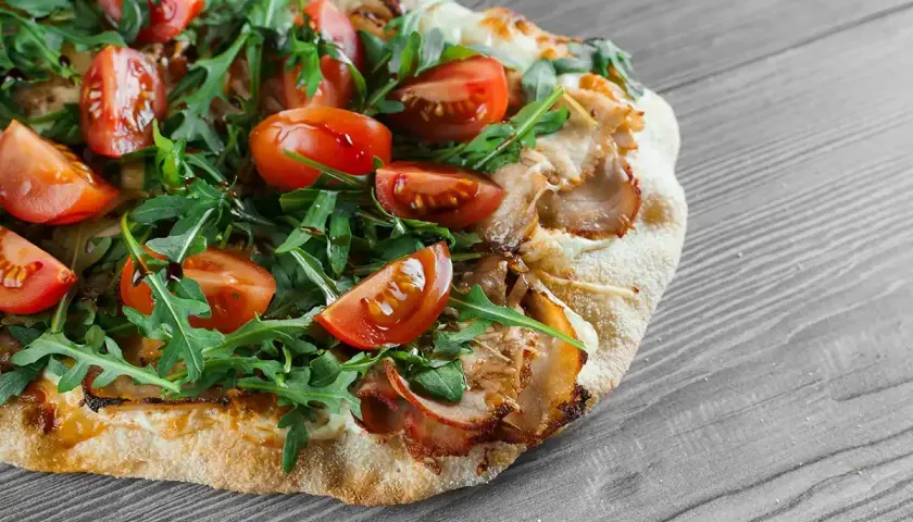 Une délicieuse pinsa recette garnie de tomates cerises, roquette fraîche et viande effilochée, sur fond de bois gris.