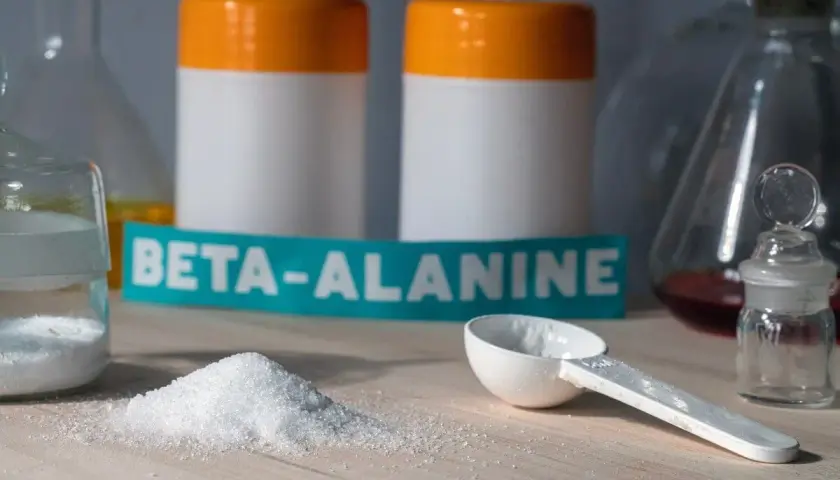 Biały proszek beta-alaniny na blacie, obok miarki i słoiczka. W tle dwa białe pojemniki z pomarańczowymi nakrętkami. Czy beta-alanina skutki uboczne są powodem do obaw?