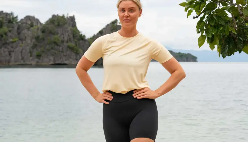 En kvinna med blont hår står på en strand med händerna på höfterna. Bakom henne syns klippor och hav. Hon ser ut som en robinsonvinnare.