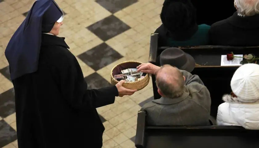 Zakonnica w czarnym habicie trzyma koszyk z pieniędzmi, do którego starszy mężczyzna wkłada banknot.