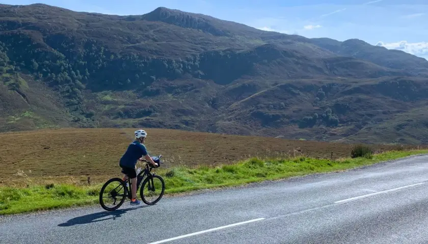 A lone cyclist enjoys a scenic bike tour Scotland, with rolling hills and mountains in the background under a bright blue sky.