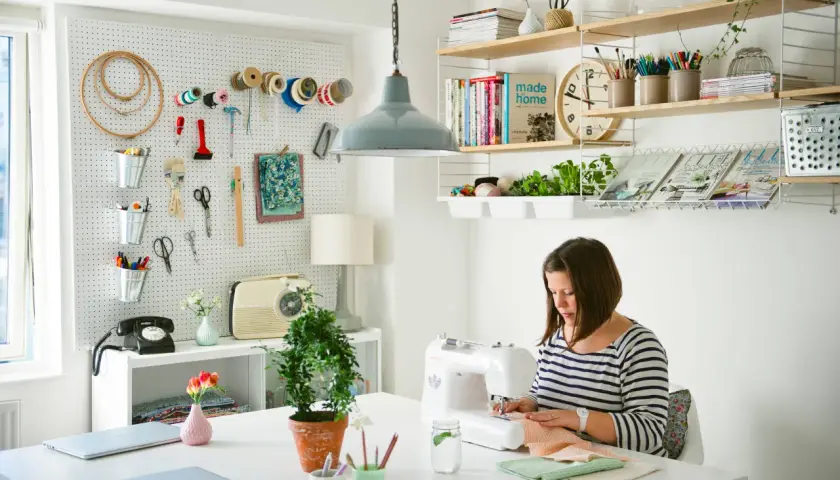 Mujer cosiendo en su **taller de manualidades en casa**. Pared con herramientas, estanterías con libros y plantas.
