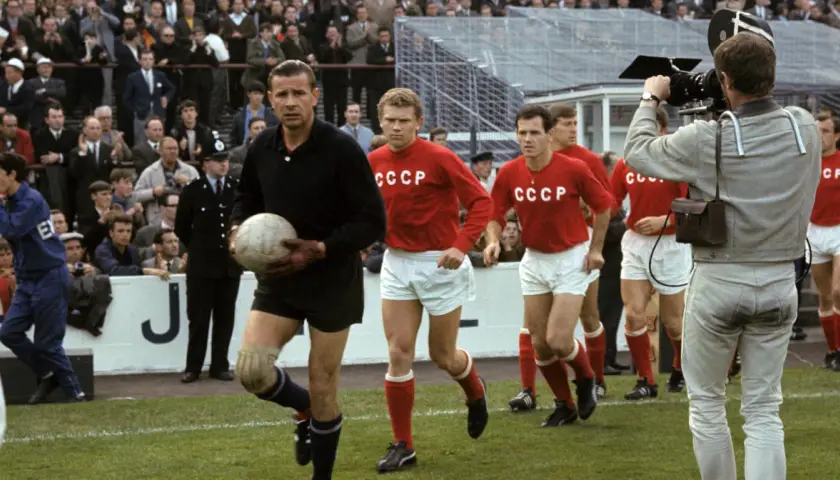 Bramkarz w czarnym stroju trzyma piłkę, obok niego biegną zawodnicy w czerwonych koszulkach z napisem CCCP. Kamera filmowa rejestruje mecz.