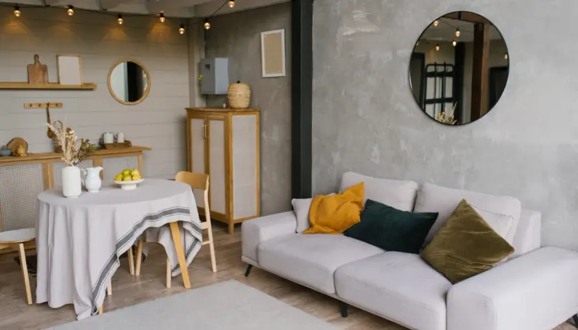 Un salon moderne avec un canapé clair, des coussins colorés et un miroir rond. La table ronde couverte d'une nappe grise et les guirlandes lumineuses ajoutent une touche chaleureuse, reflétant une nouvelle tendance déco.