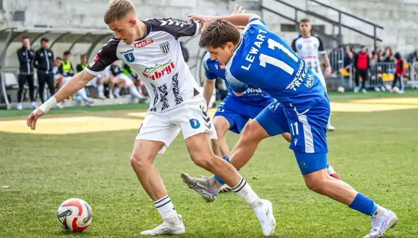 Piłkarze walczą o piłkę na boisku. Jeden w białej koszulce, drugi w niebieskiej z numerem 11. Emocje jak w walce o 3 liga tabele.