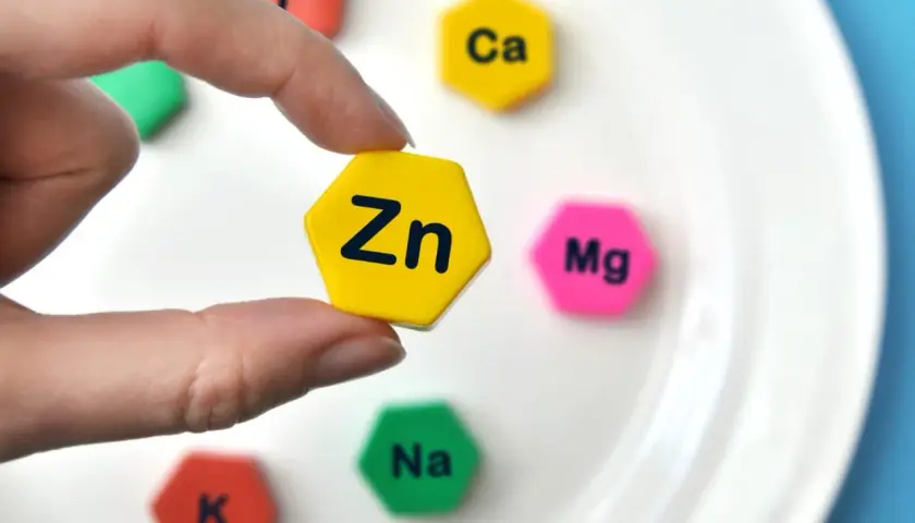Palec wskazujący trzyma żółty heksagon z napisem "Zn". Wokół niego na białym talerzu leżą inne heksagony z symbolami pierwiastków: Ca, Mg, Na, K.