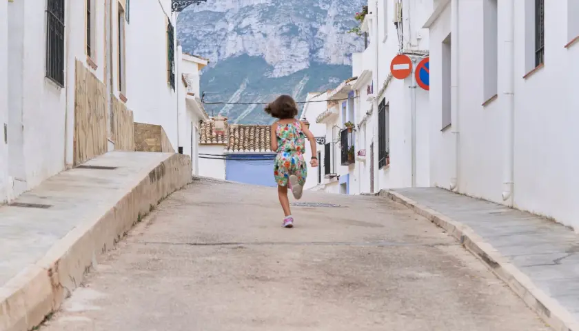 Niña corre por calle empinada de un pueblo de Alicante, con casas blancas y montañas al fondo.