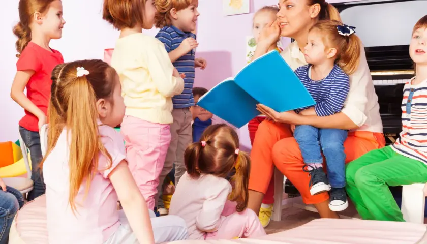 Nauczycielka czyta dzieciom książkę, pokazując różne rodzaje zajęć w przedszkolu. Dzieci słuchają z uwagą.