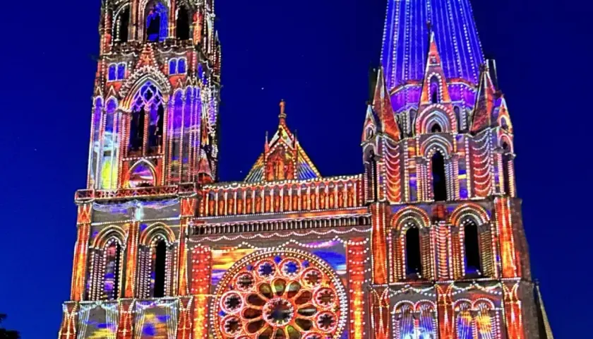 Katedra w Chartres nocą, rozświetlona kolorowymi projekcjami. Architektura gotycka w odcieniach niebieskiego i pomarańczowego.