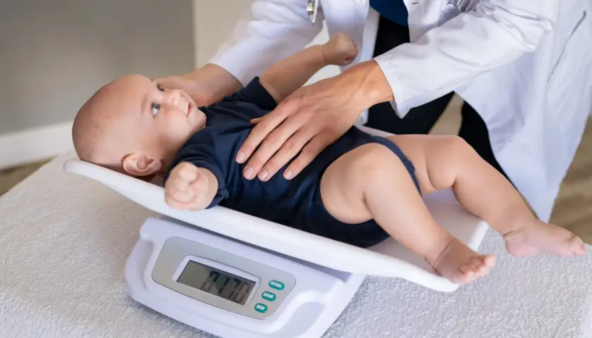 Bebé de 11 meses en báscula digital, con manos de doctor. El peso del bebé es de 9.70 kg.
