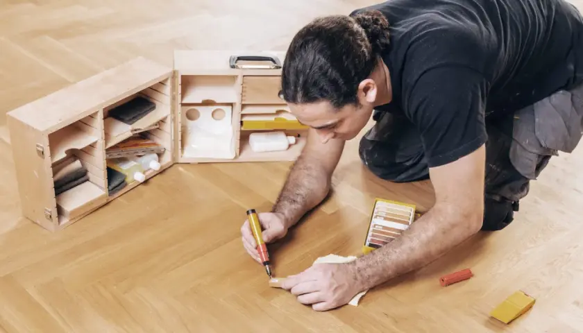 Mann repariert Parkett mit einem Reparaturstift und einer Farbkarte. Werkzeugkasten daneben.