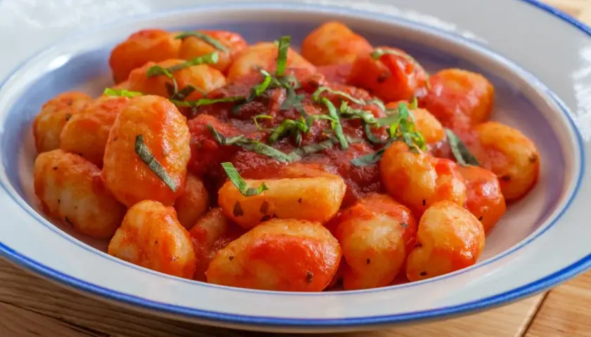Pyszne gnocchi z sosem pomidorowym i świeżą bazylią, podane w białej misce z niebieskim rantem.