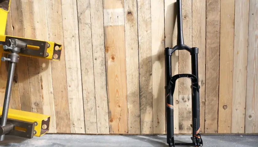 A black MRP Ribbon coil suspension fork stands against a wooden wall. The fork features orange and grey accents and the "ribbon" branding is visible.