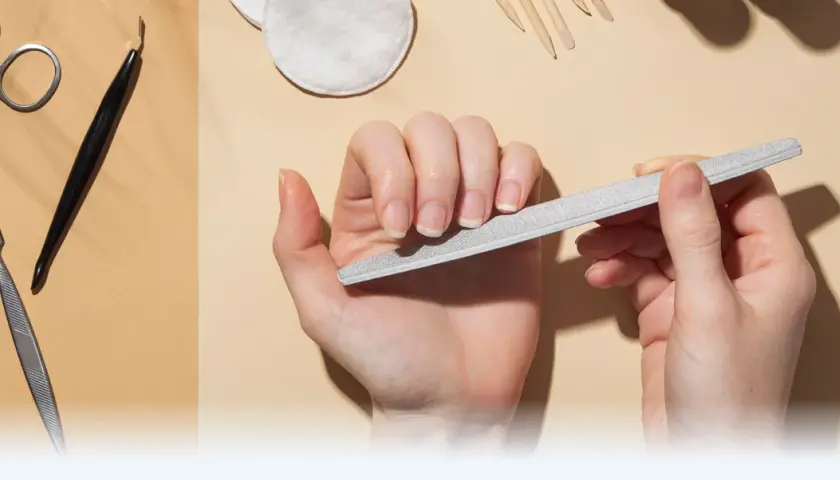 Dłonie pielęgnują paznokcie, używając pilnika. W tle widać narzędzia do manicure, takie jak nożyczki i cążki, tworząc kompletny zestaw do pielęgnacji macierzy paznokcia.