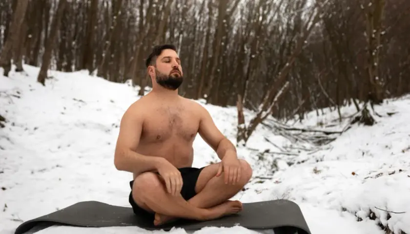 Mann in der Natur, barbusig im Schnee sitzend, praktiziert Wim Hof Atmung.
